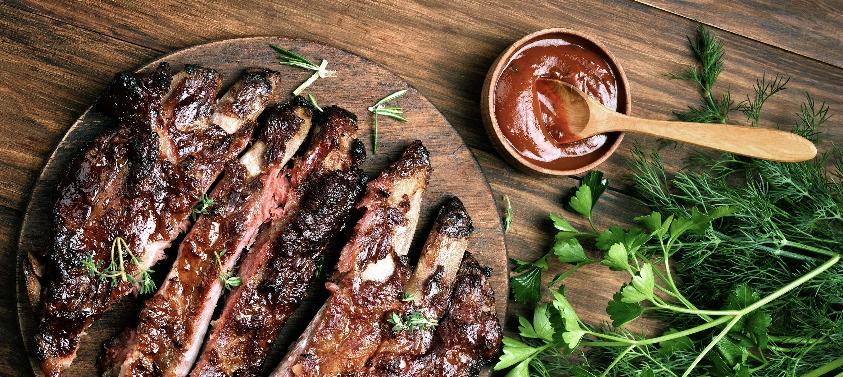 Barbecue ribs on a plate with fresh green herbs and a wooden spoon resting in a cup of BBQ sauce. A bold, summer-ready grilling image.