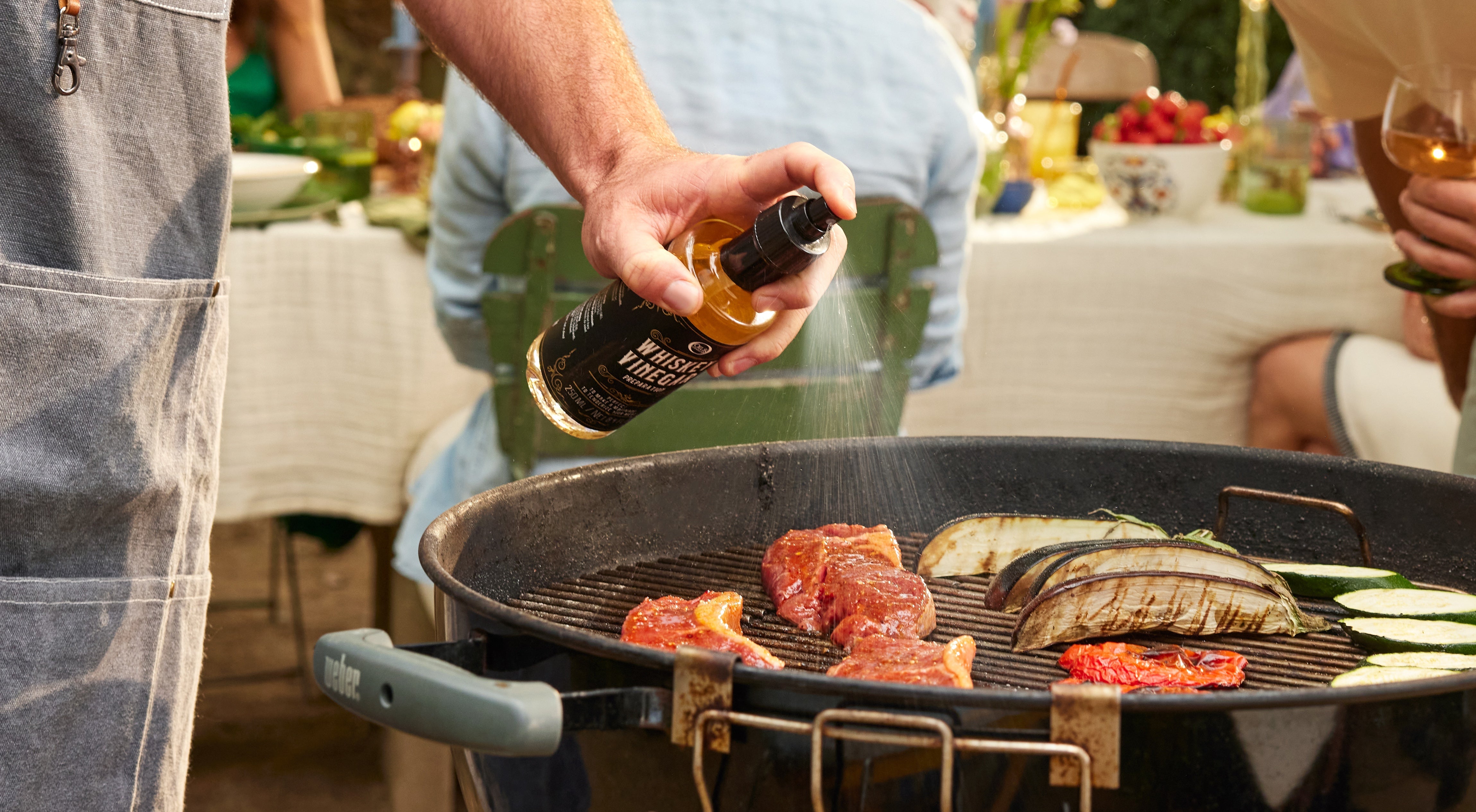 Backyard BBQ scene with someone pouring olive oil onto food on the grill — capturing the flavor and fun of specialty oils and vinegars.